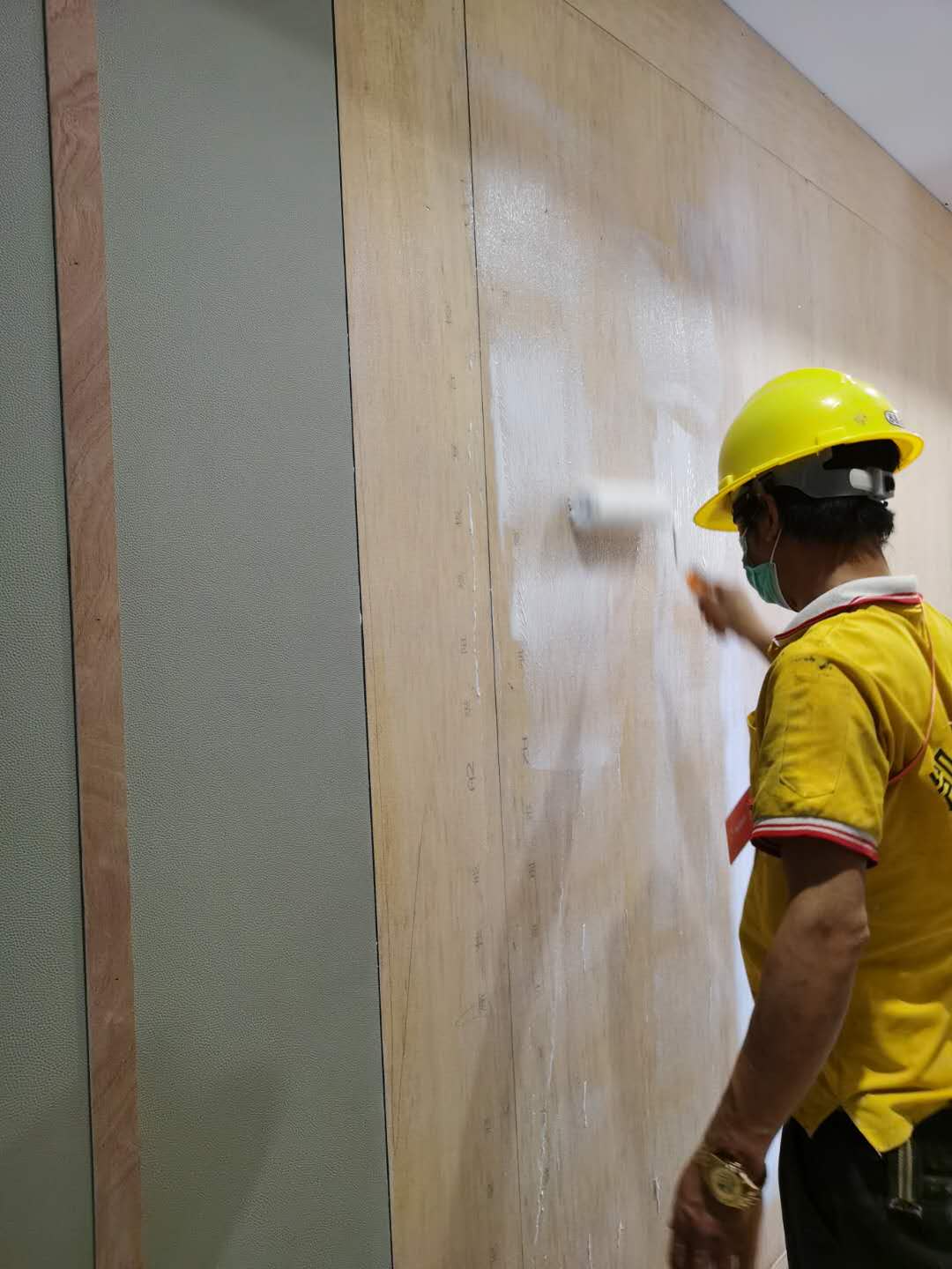 工人戴安全帽用滚筒在墙面基层滚涂胶粘剂，防撞软包安装施工现场，适用于办案区审讯室软包装修